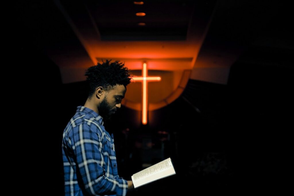 A man in a plaid shirt stands indoors reading a book, with a large illuminated cross in the background.