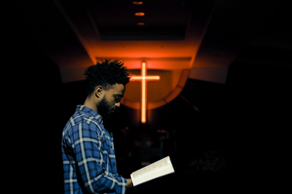 A man in a plaid shirt stands indoors reading a book, with a large illuminated cross in the background.