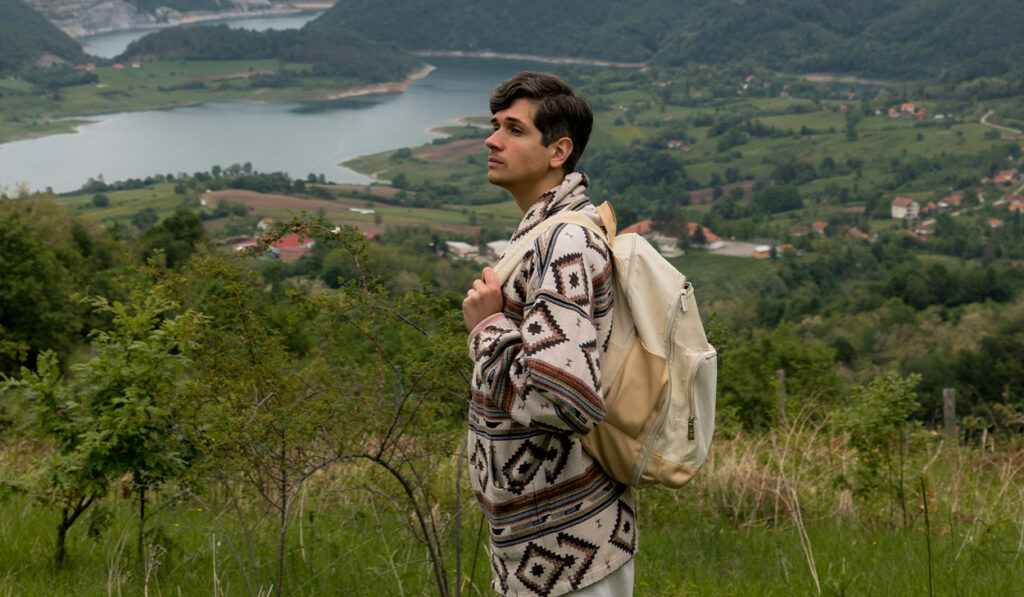 A man with a backpack stands in front of green scenery.