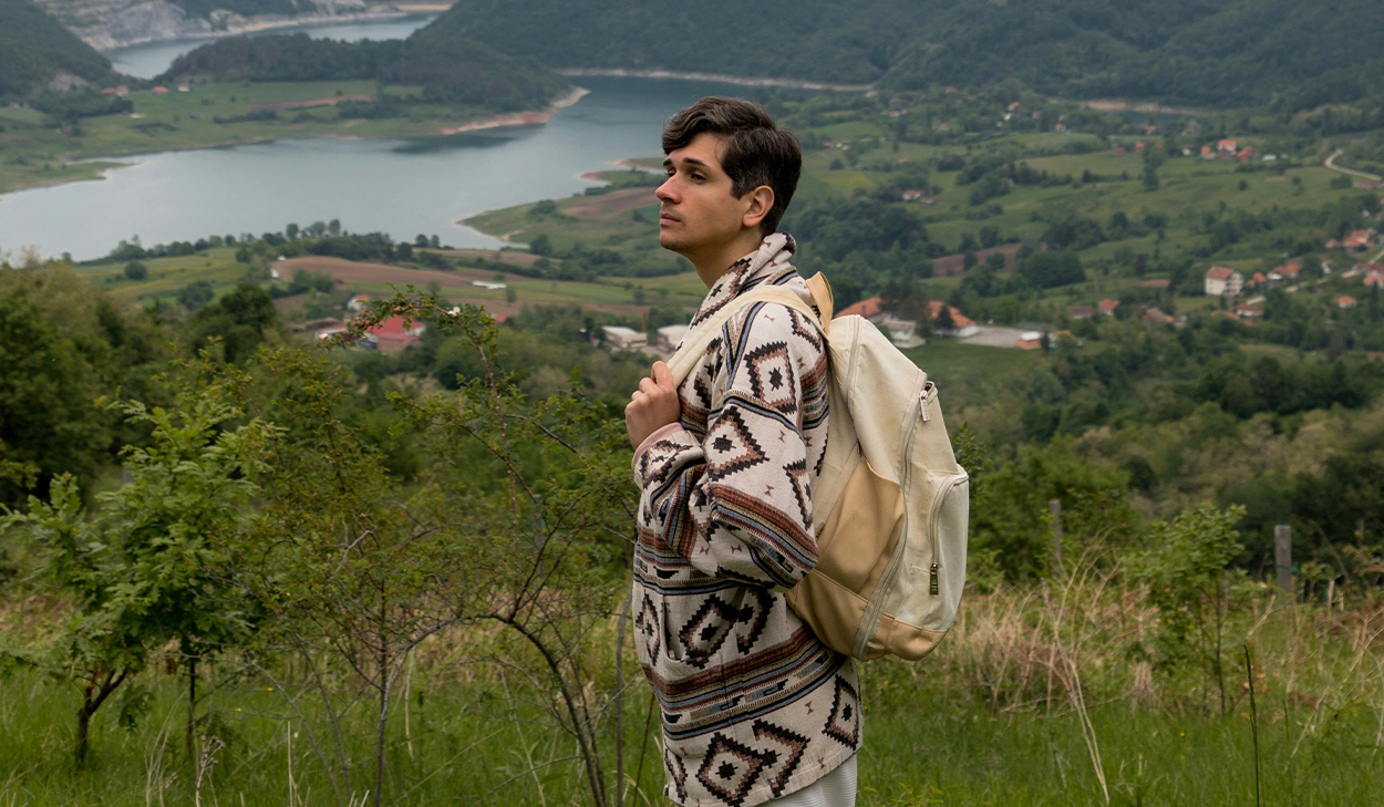 A man with a backpack stands in front of green scenery.