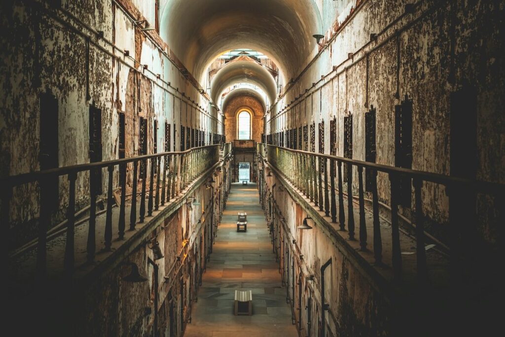 Long, narrow corridor in an old, rundown prison with rows of cell doors on both sides, peeling walls, and an arched ceiling illuminated by natural light at the end.