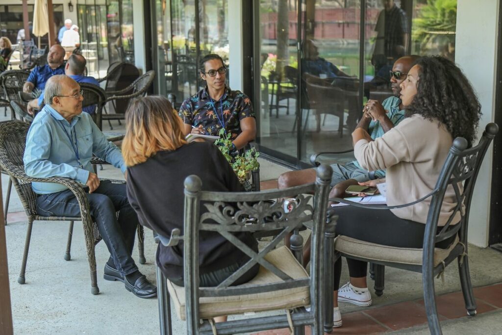 Five people sit in a circle outdoors, engaged in conversation near a building with large windows and patio furniture.