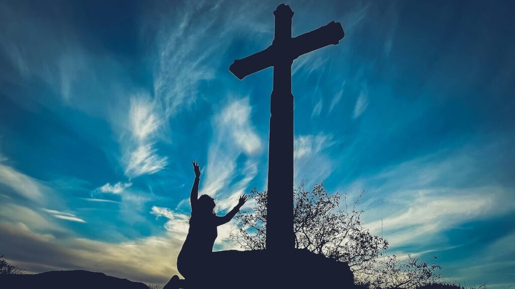 Silhouette of a person with raised arms kneeling beside a large cross outdoors at sunset, with dramatic clouds in the sky.