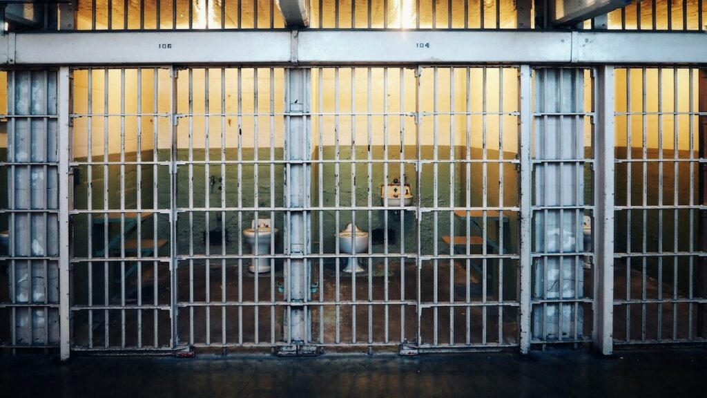 A row of empty prison cells behind metal bars, with yellow lighting inside each cell and a walkway in front.