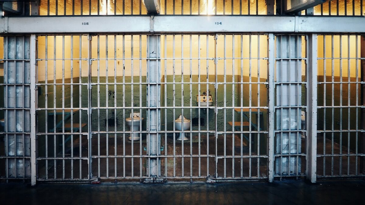 A row of empty prison cells behind metal bars, with yellow lighting inside each cell and a walkway in front.