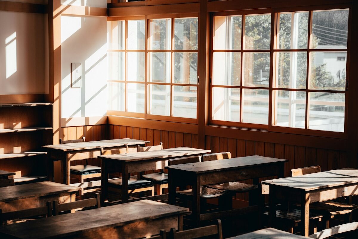Sunlight streams through large windows into an empty classroom with wooden desks and chairs arranged in rows.