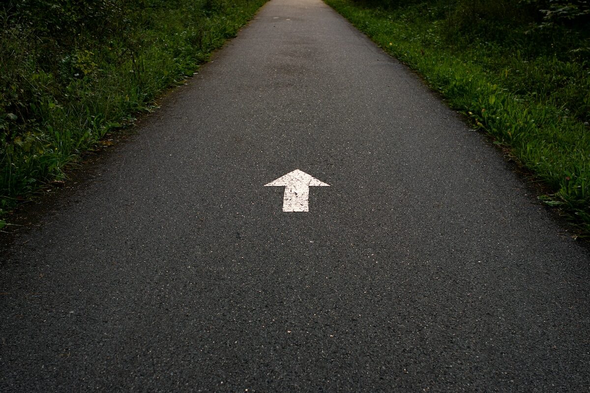 A paved path with a white arrow painted in the center, pointing straight ahead, bordered by grass on both sides.