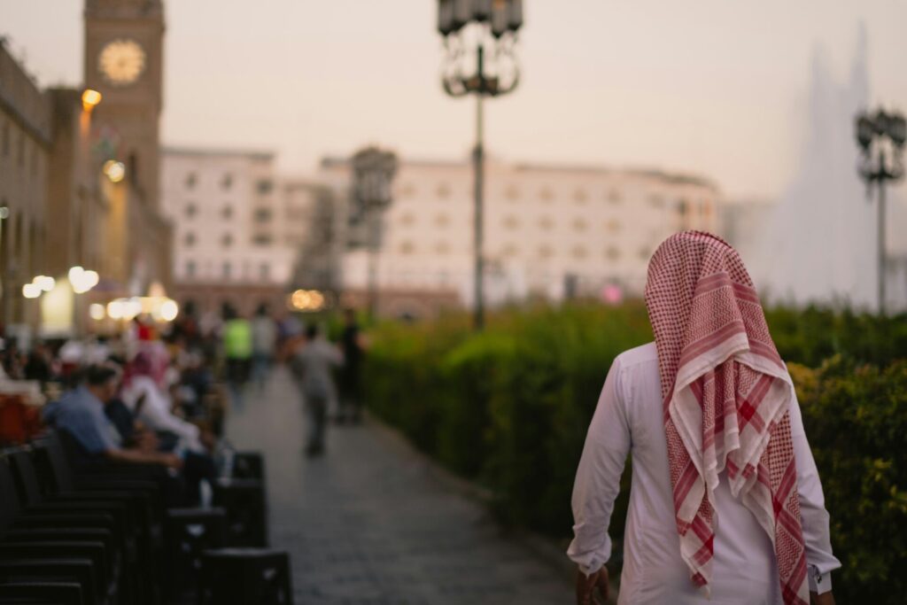 A man in a red and white headscarf walking away from the camera on sidewalk during sunset