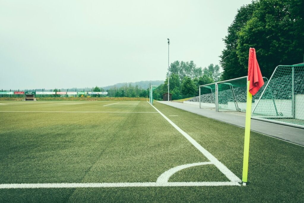 A soccer field corner with a yellow flagpost and red flag, empty stands, and trees in the background under a cloudy sky.