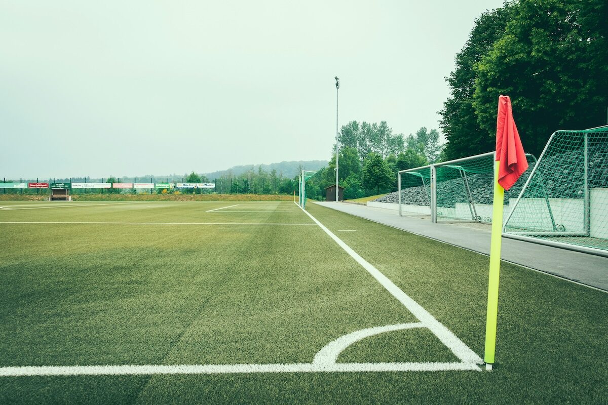 A soccer field corner with a yellow flagpost and red flag, empty stands, and trees in the background under a cloudy sky.