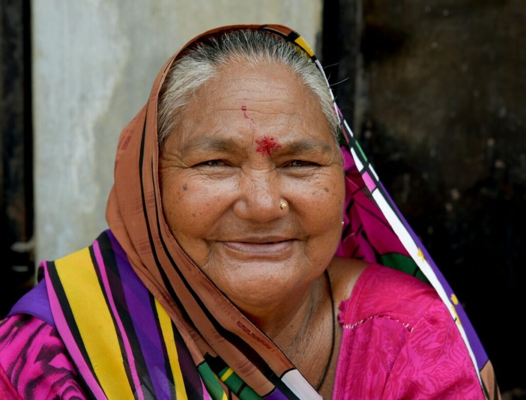 Elderly woman with gray hair smiles at the camera, wearing a colorful sari with orange, purple, and yellow patterns, and a red bindi on her forehead.