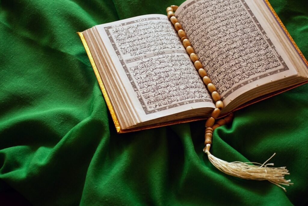An open Quran with prayer beads placed on its pages, resting on a green fabric.