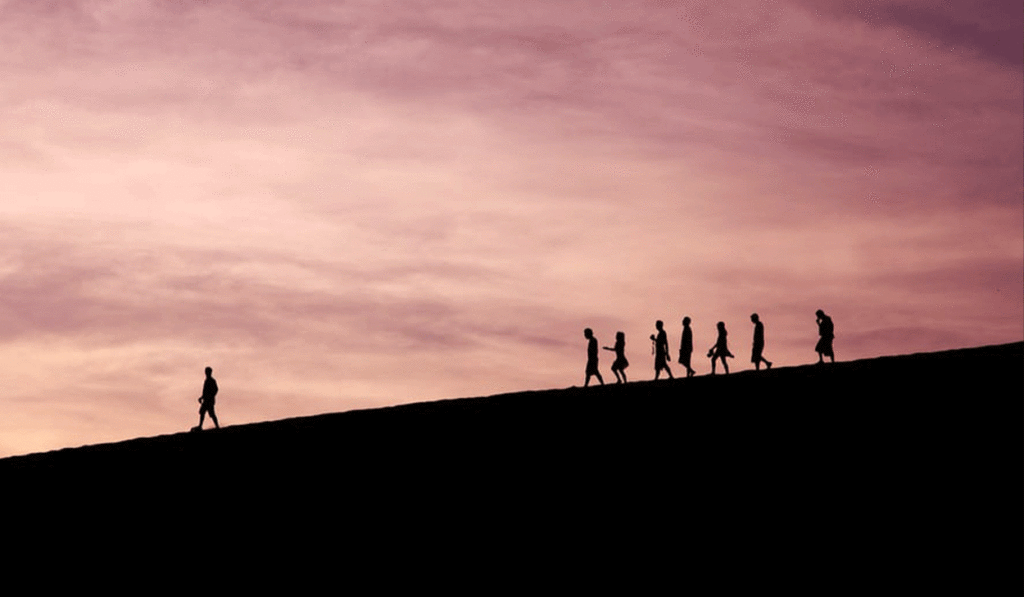 Silhouettes of seven people walking in a line on a hill at sunset, with one person walking ahead of the group.