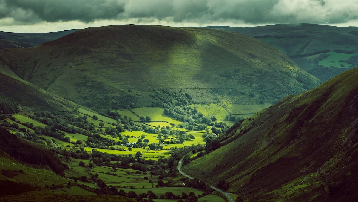 A green valley with scattered trees and fields is surrounded by large hills under a cloudy sky, with sunlight creating patches of light on the landscape.