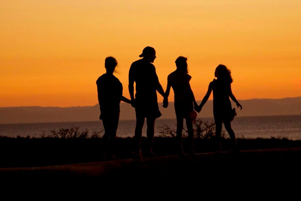 Four people are holding hands and walking outdoors at sunset, silhouetted against an orange sky.