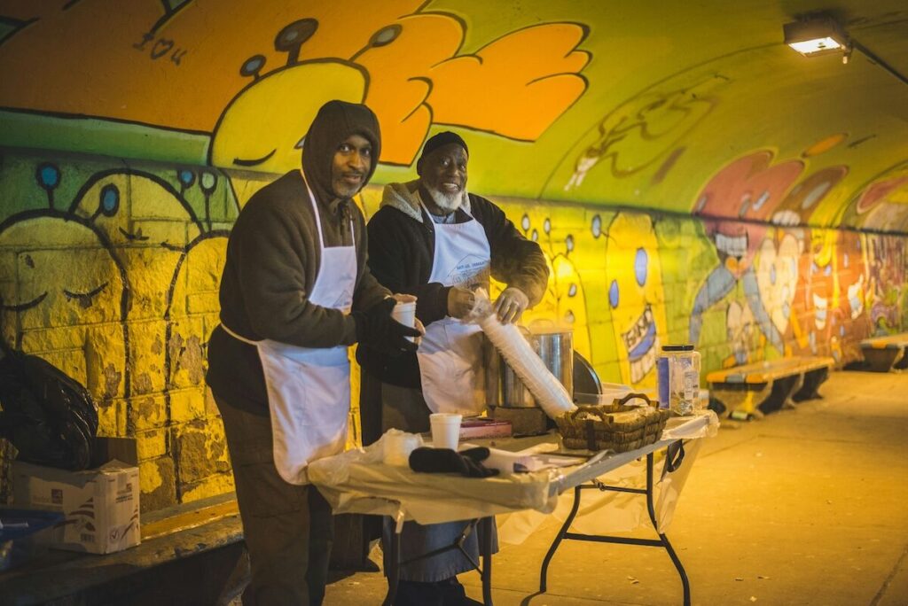 Two people wearing aprons serve food at a table in a brightly painted, graffiti-covered tunnel. Supplies and food items are laid out on the table.