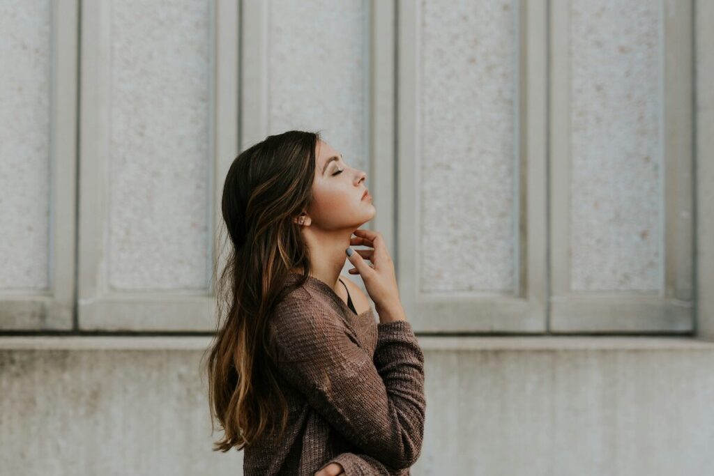 A young woman with long brown hair stands in profile against a textured wall, looking upward with her hand near her neck.