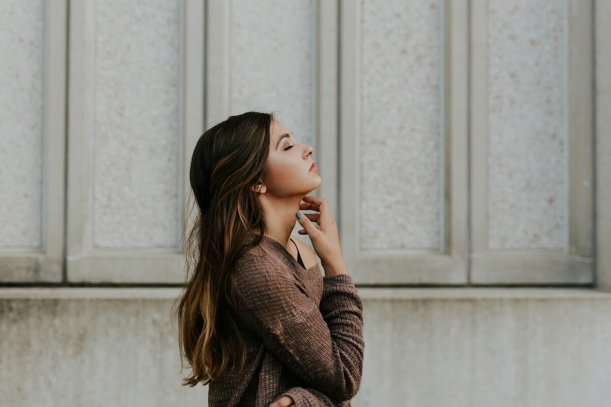 A young woman with long brown hair stands in profile against a textured wall, looking upward with her hand near her neck.