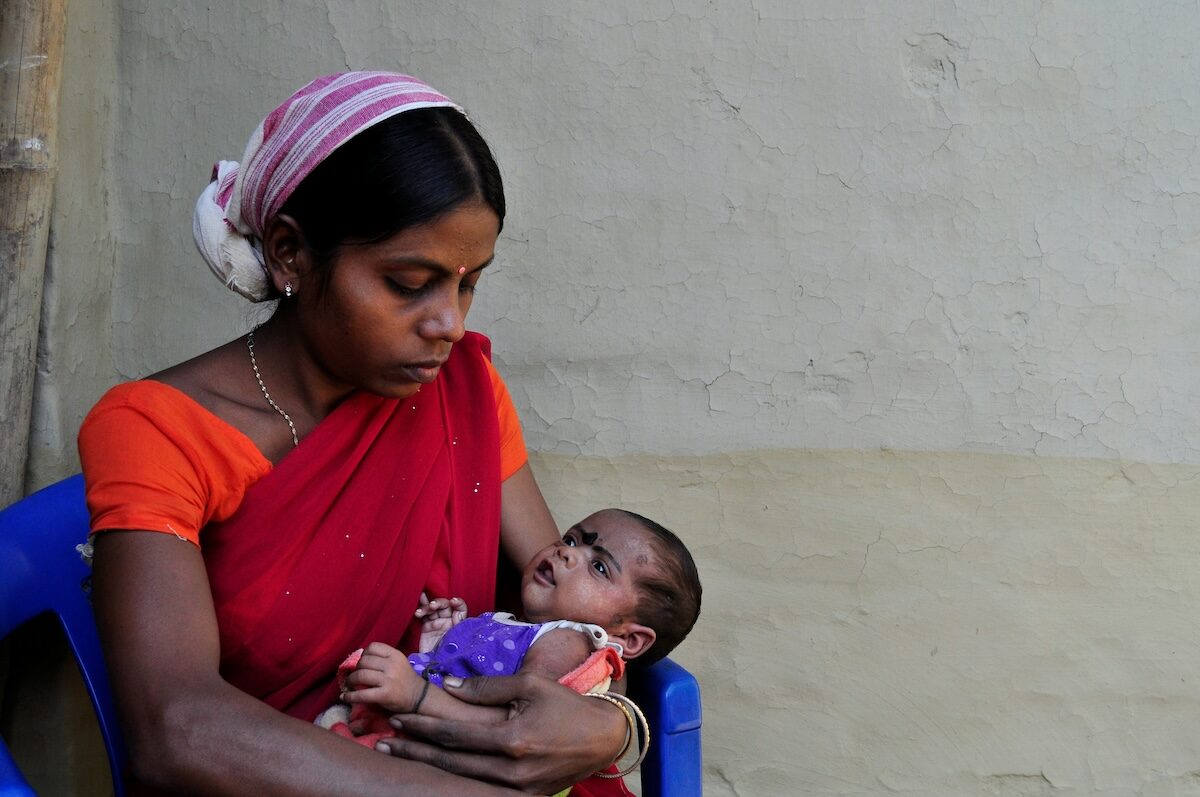 A woman in a red sari sits on a blue chair, holding a baby in her arms while looking down at the child.