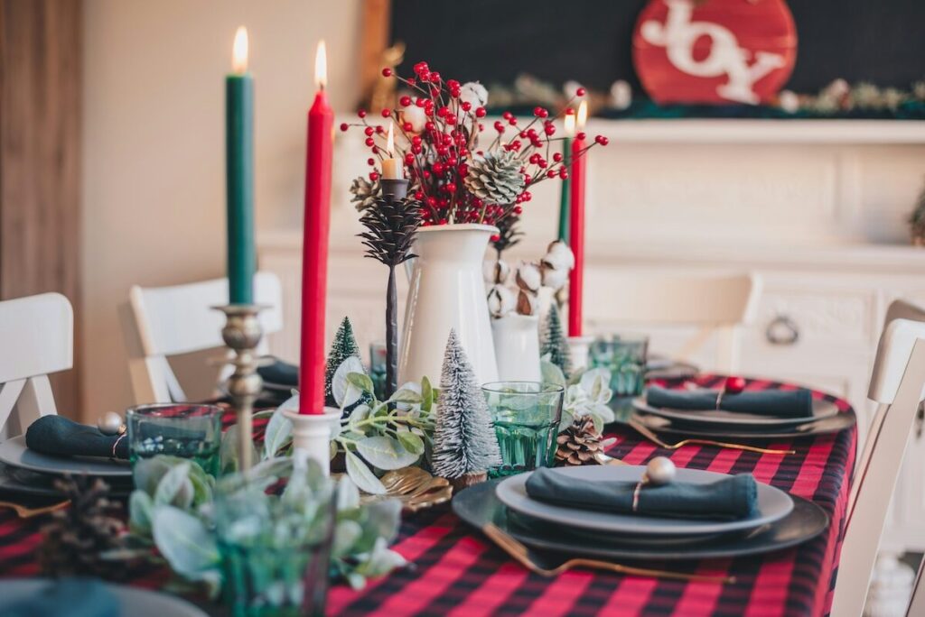 A dining table set for a holiday meal features black plates, green glasses, red and green candles, a plaid tablecloth, and festive centerpiece with greenery and berries.