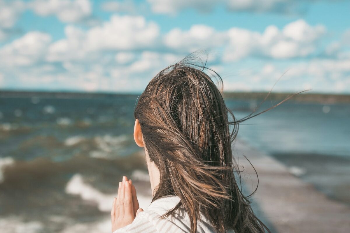 Person with long brown hair stands facing the ocean with hands together, under a partly cloudy sky.