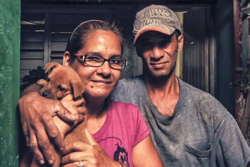 A woman in glasses holds a small brown puppy while standing next to a man in a cap and t-shirt. Both are looking at the camera and smiling slightly.
