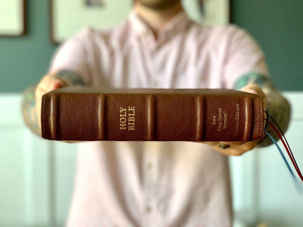 A person in a light pink shirt holds out a closed Holy Bible with a brown leather cover and gold lettering on the spine.