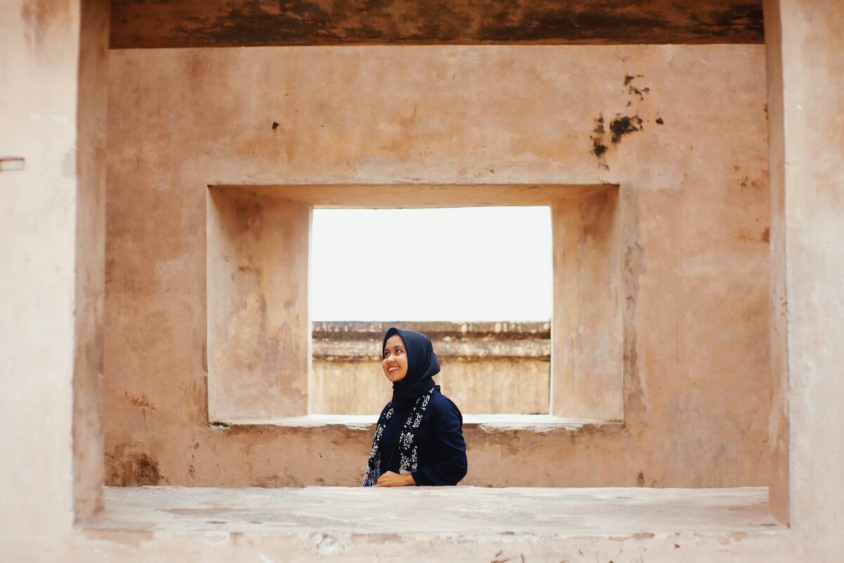 A person wearing a dark headscarf stands in front of a rectangular window in an aged, beige stone wall.