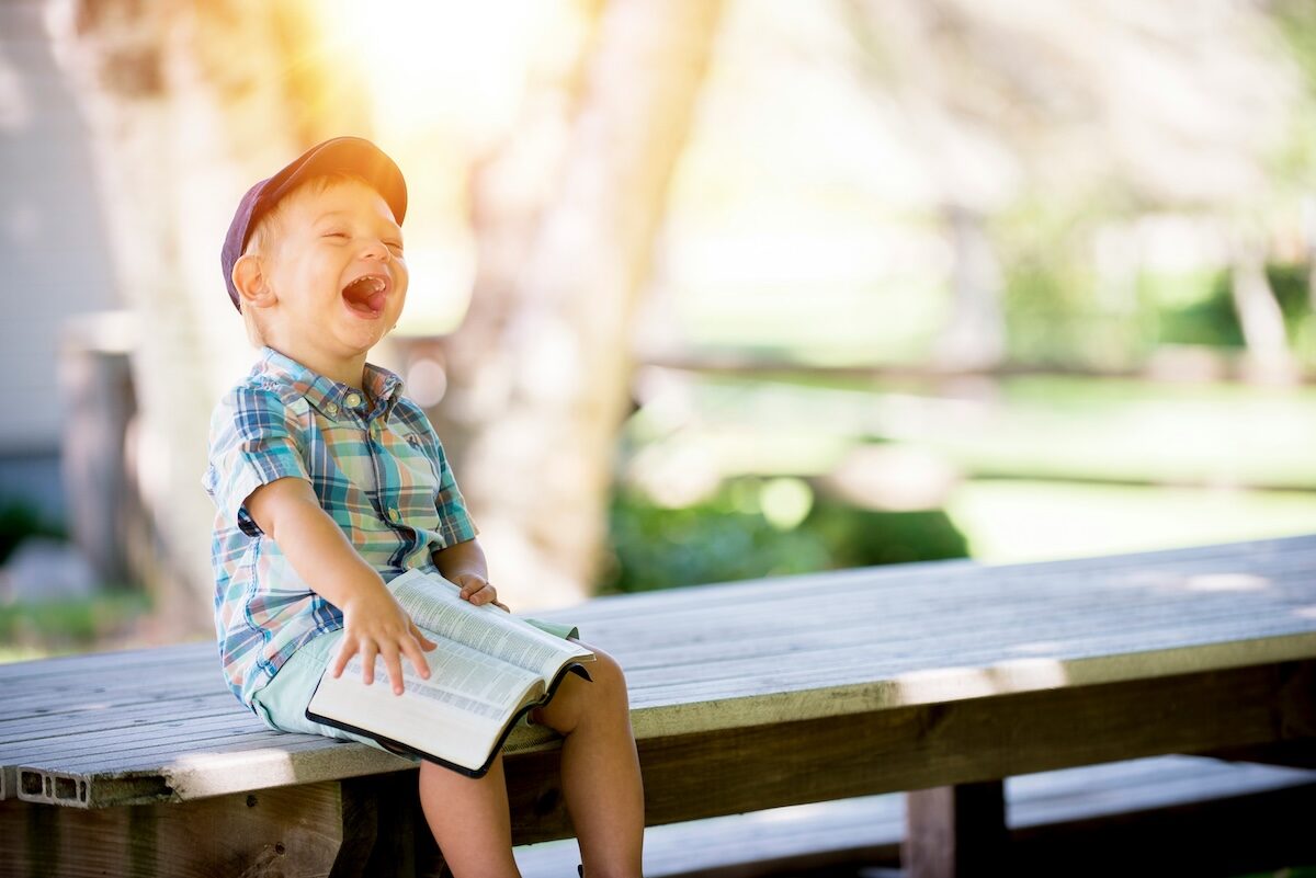 A young child wearing a cap and plaid shirt sits on a wooden bench outdoors, laughing while holding an open book in their lap.