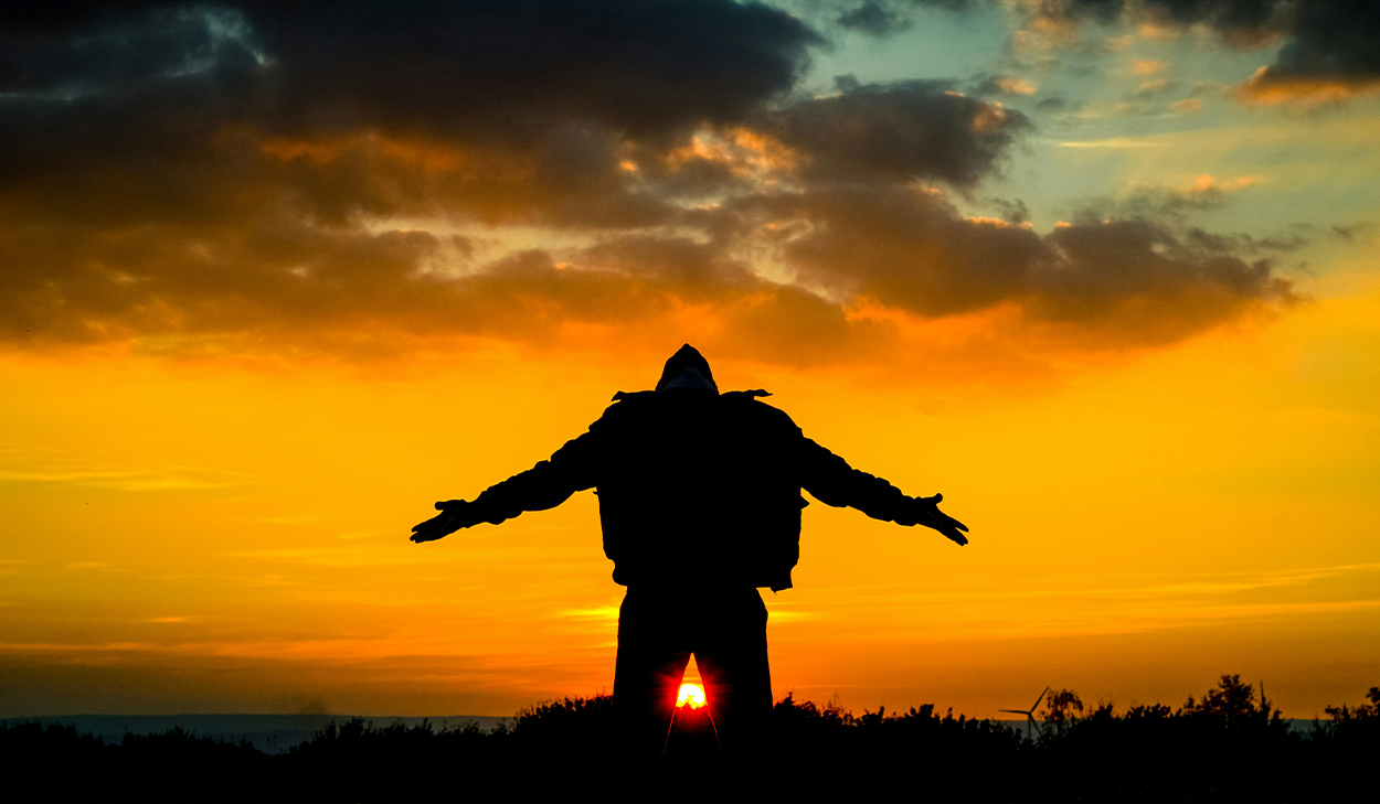 A man with a hooded jacket stands with his arms outstretched. He is backlit by the setting sun.