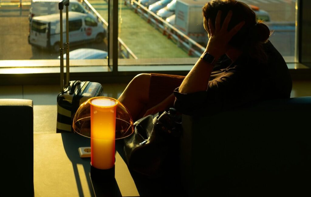 A person sits in a dimly lit airport lounge with a suitcase and bag, resting their head on their hand near a glowing lamp. Cars are visible through the window outside.