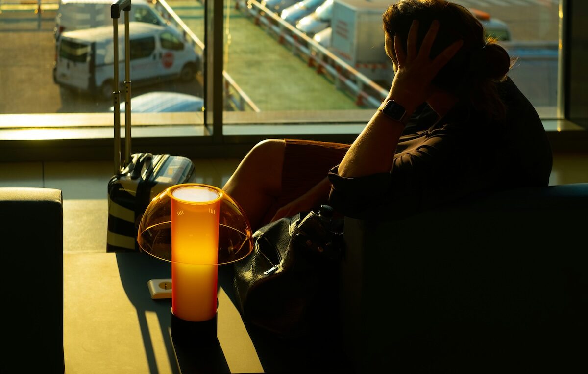 A person sits in a dimly lit airport lounge with a suitcase and bag, resting their head on their hand near a glowing lamp. Cars are visible through the window outside.