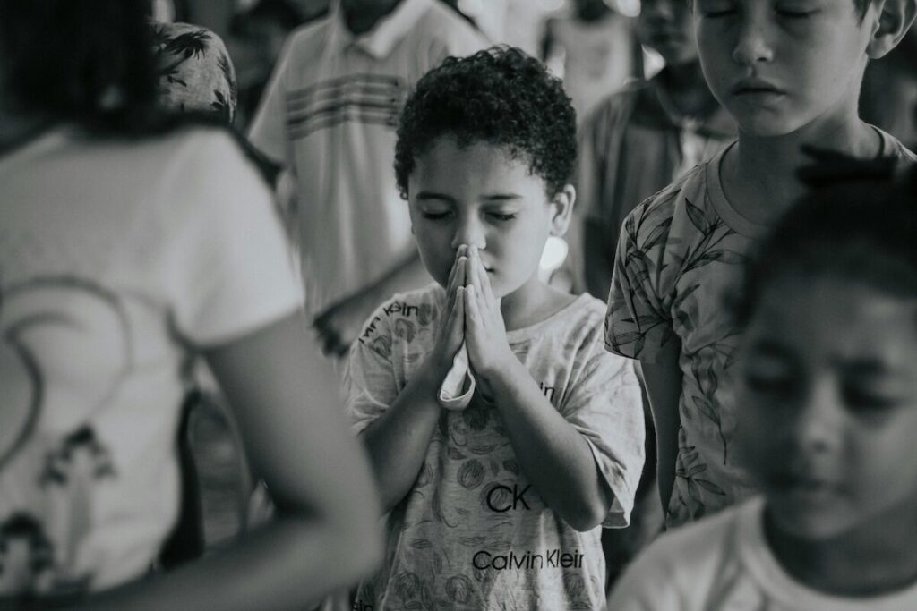 A young child stands with eyes closed and hands pressed together in prayer, surrounded by other children, in a black and white photo.