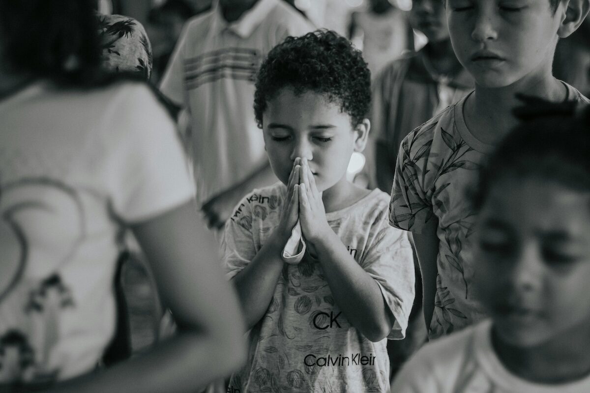 A young child stands with eyes closed and hands pressed together in prayer, surrounded by other children, in a black and white photo.
