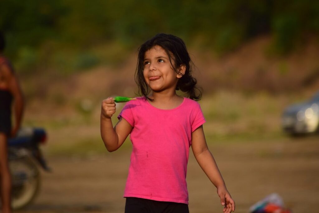 A young girl in a pink shirt stands outdoors, smiling and holding a green object, with a blurred background of trees and a motorcycle.
