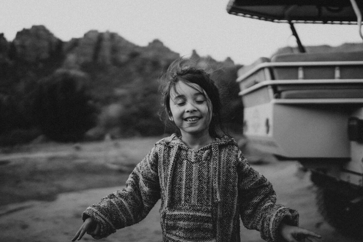 A child stands outdoors near a vehicle, smiling with eyes closed. Rocky hills and trees are visible in the background. The image is in black and white.