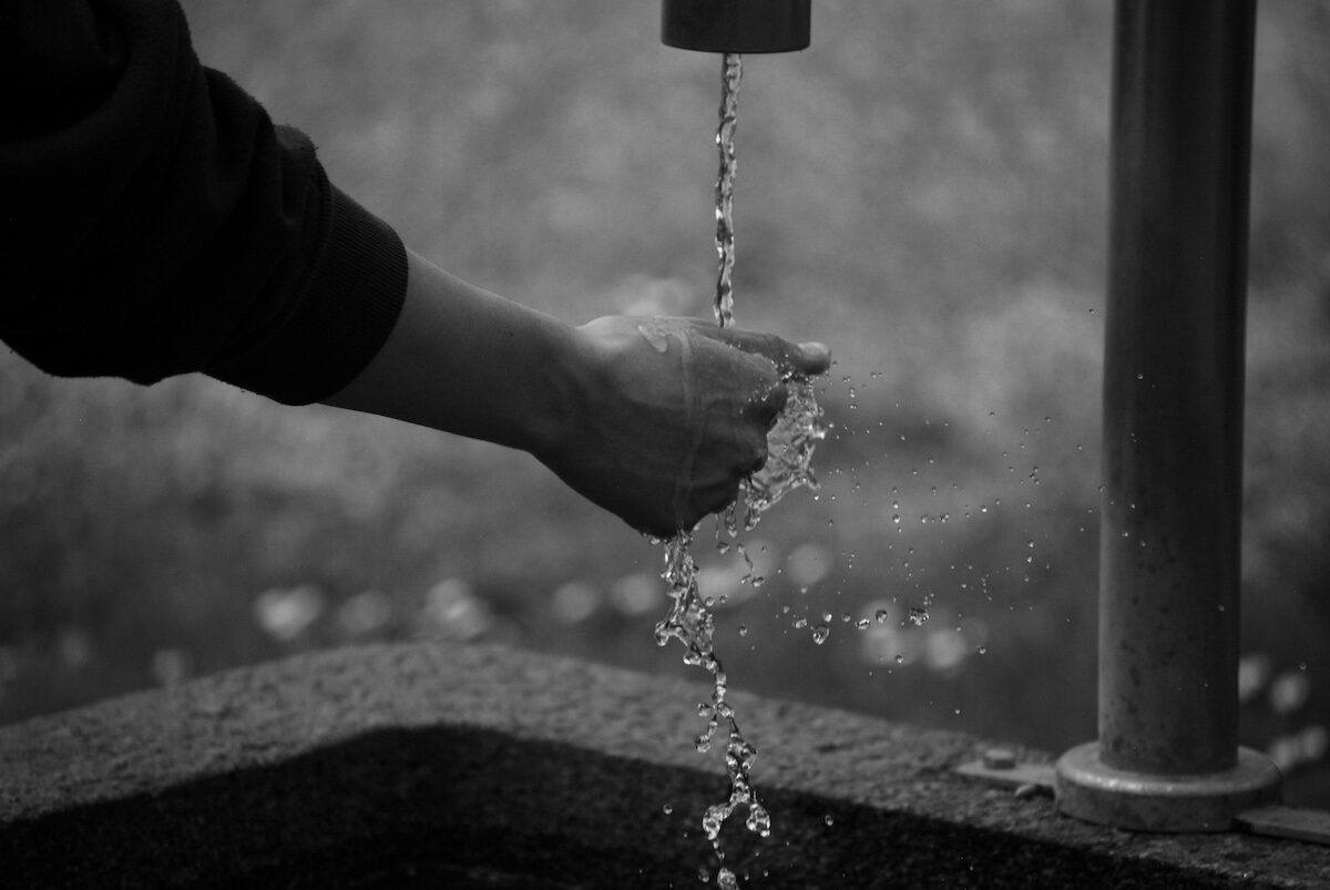 A person’s hand is held under running water from a faucet, with water splashing into a basin below. The image is in black and white.
