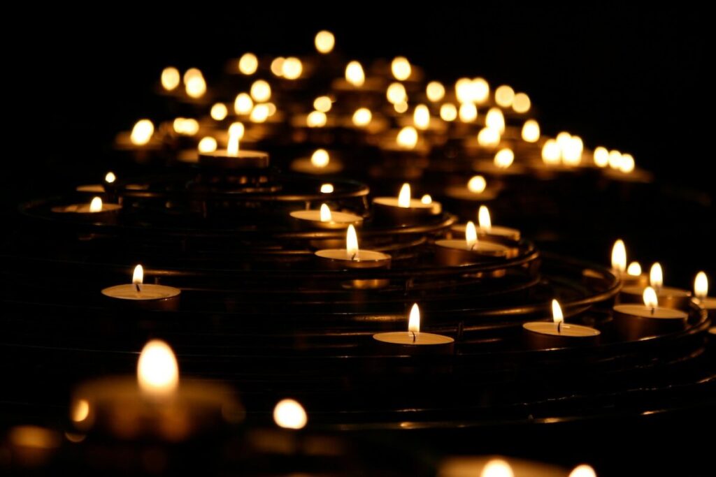Dozens of small tea light candles are lit and arranged in rows, creating a warm glow against a dark background.