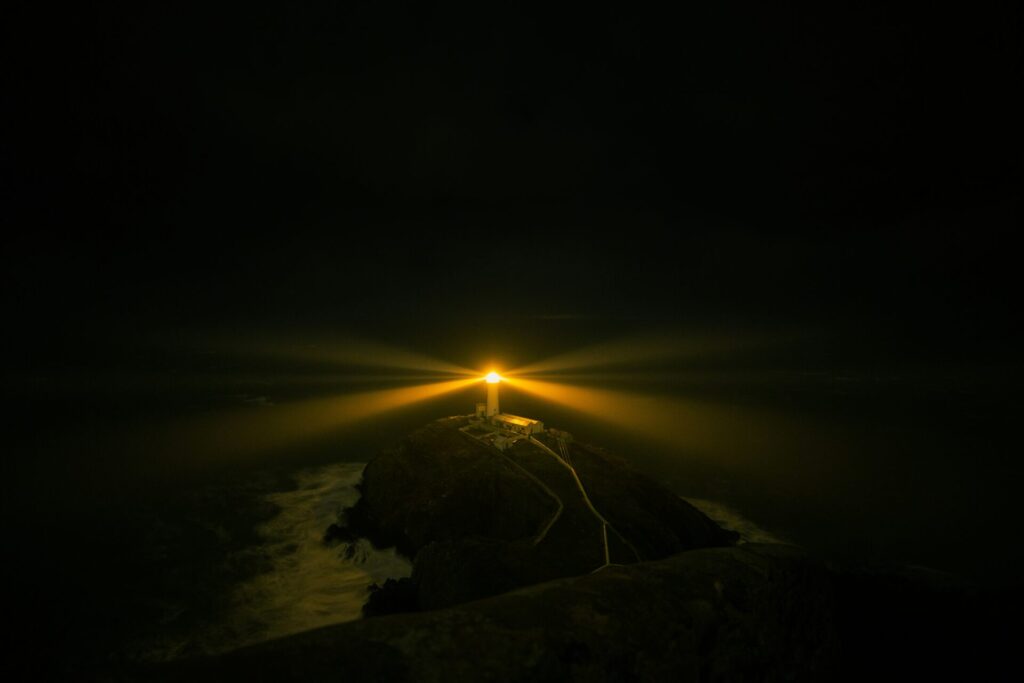 A lighthouse on a rocky coast emits a bright beam of light, illuminating the surrounding darkness at night.