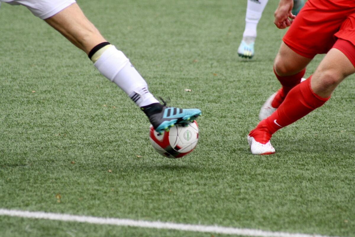 Two soccer players, one in white and the other in red, compete for the ball on a grass field; only their legs and feet are visible.