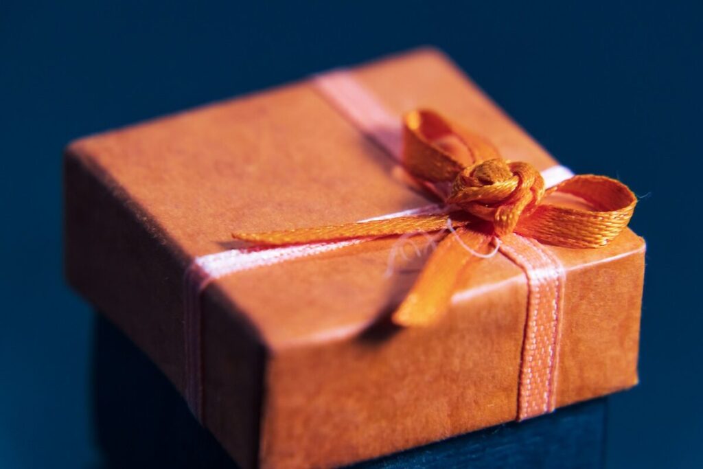 A small, square gift box wrapped in orange paper with a matching orange ribbon tied in a bow, set against a dark blue background.