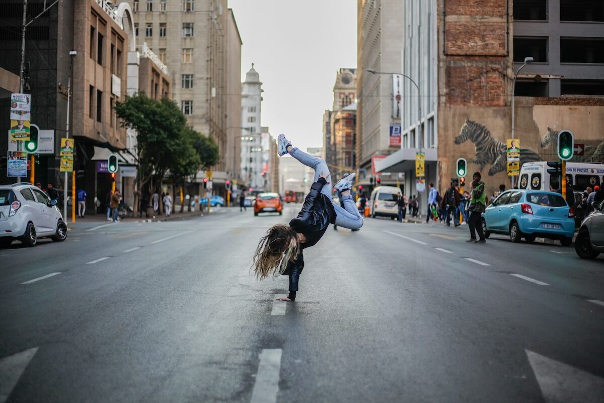 A person performs a one-handed breakdance move in the middle of a city street, surrounded by cars and tall buildings.