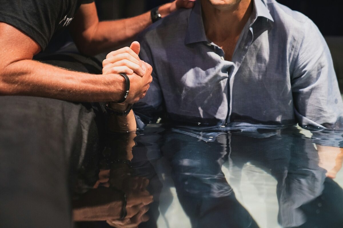 Two people are partially submerged in water; one person is gripping the other’s hand, suggesting a supportive or ceremonial moment.