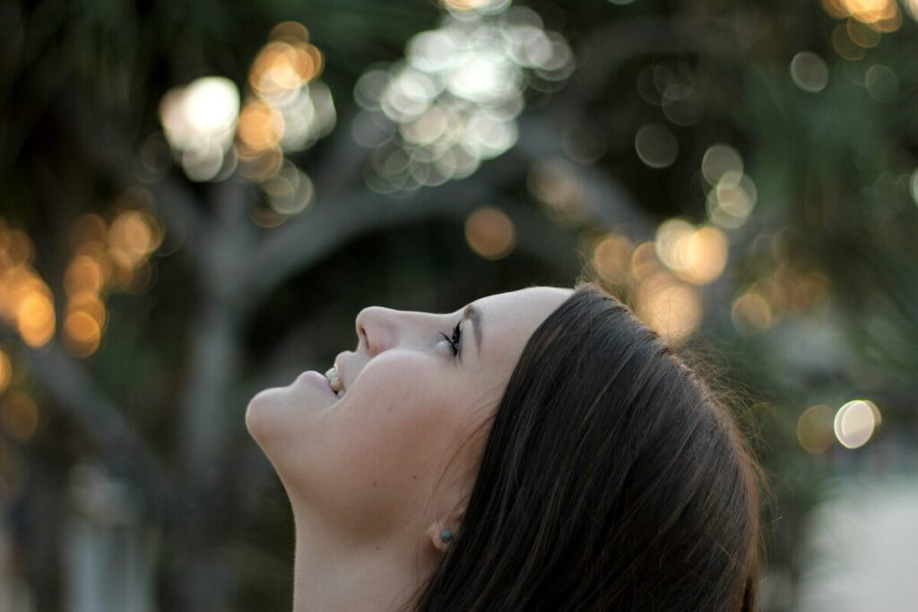 A woman with long brown hair looks upward, with a soft background of blurred trees and sunlight.