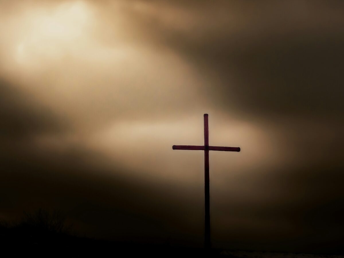 A simple wooden cross stands silhouetted against a cloudy sky with light shining through the clouds.