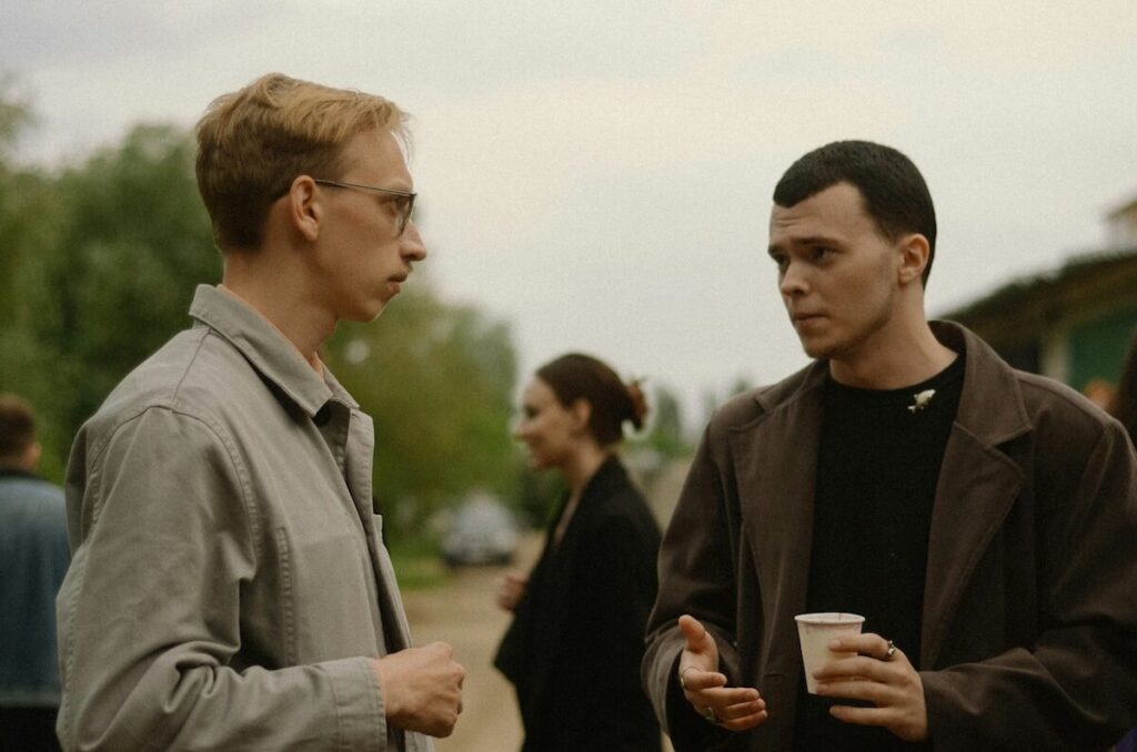 Two men stand outdoors talking; one holds a paper cup while gesturing, and a woman is visible in the blurred background.