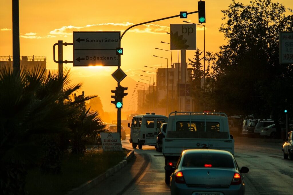Cars driving on a city street at sunset with traffic lights illuminated and road signs visible in the background.