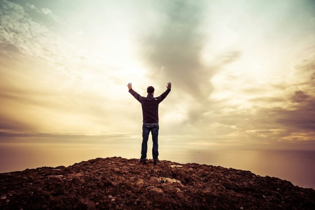 A person stands on a rocky hilltop with arms raised, facing a bright cloudy sky at sunrise or sunset.