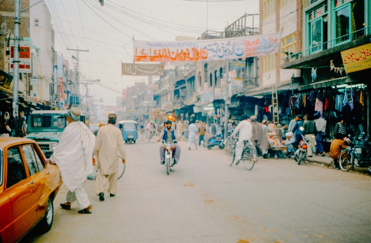 A busy street scene with people walking, cyclists, cars, and shops lining both sides of the road, and a large banner hanging overhead.