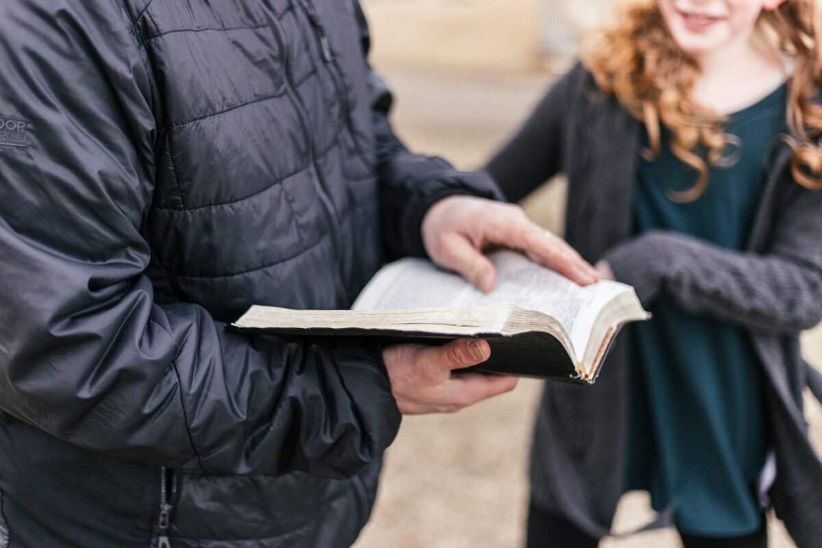 An adult wearing a dark jacket holds an open book, possibly a Bible, while a child with long curly hair stands nearby.